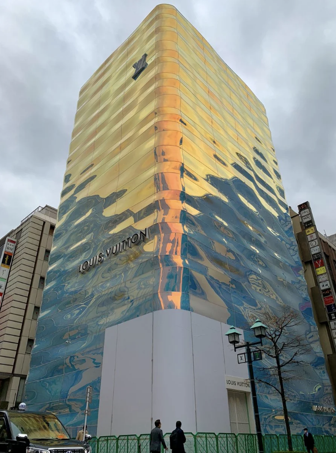 Glass building facade with reflective design and "Louis Vuitton" logos, pedestrians nearby