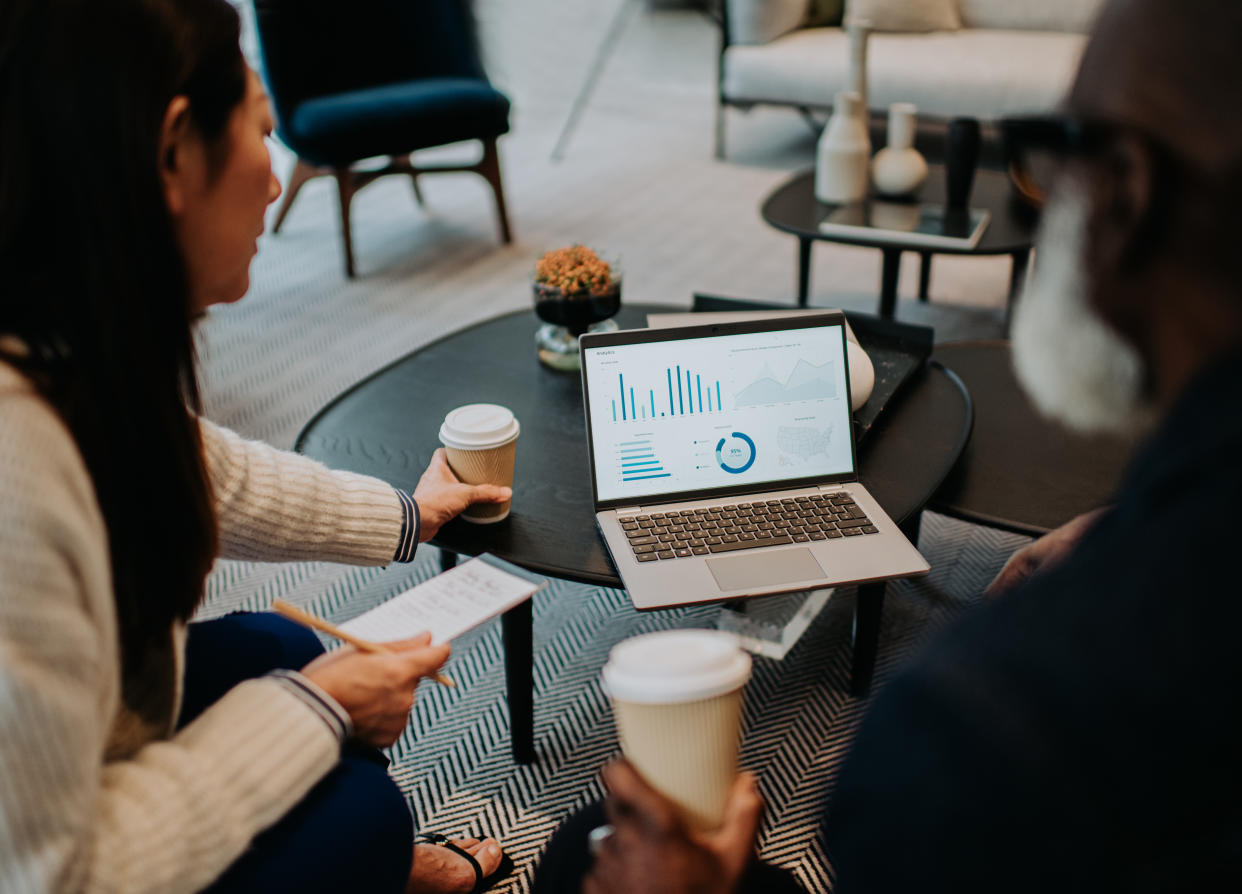 Colleagues sit on a sofa and have a casual meeting. They share a laptop, both looking at the statistics displayed, while they drink coffee.