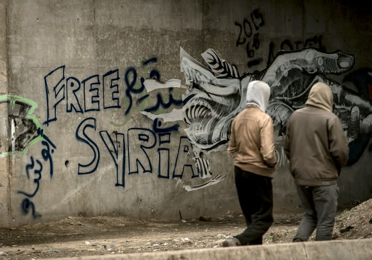 Migrants walk past graffiti reading "Free Syria" in the "Jungle" migrant camp in Calais, northern France, on February 12, 2016