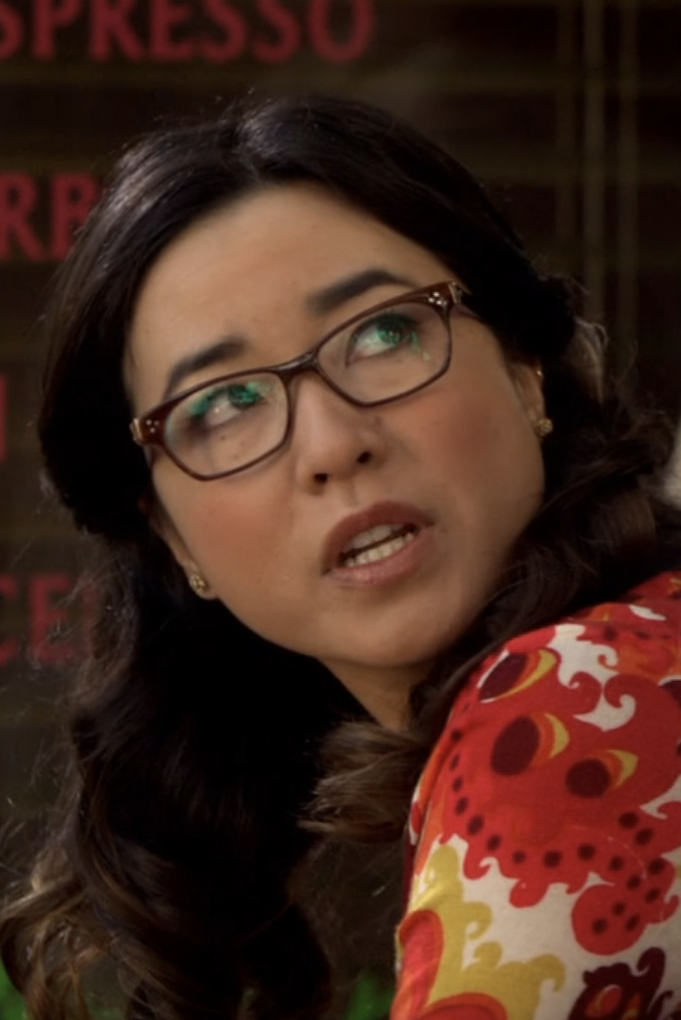 A young Maya Erskine with long dark hair and glasses looks up, wearing a red patterned outfit