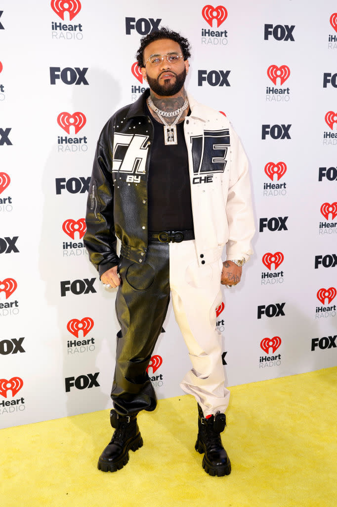 Person on yellow carpet posing in front of backdrop with logos, wearing a graphic leather jacket, pants, and chunky boots