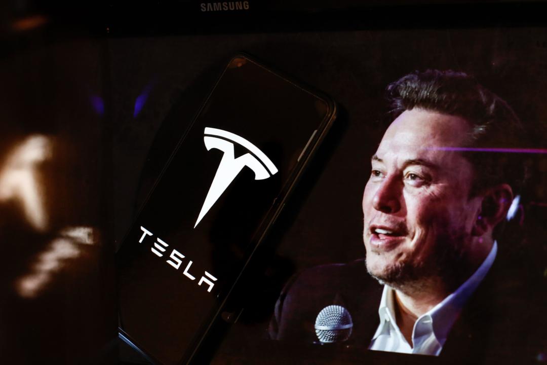 In this photo illustration, the Tesla logo is seen displayed on a smartphone screen and a computer screen displaying a portrait of Elon Musk, who is the owner of Tesla. The company recently lost clients due to Elon Musk's right wing shift.