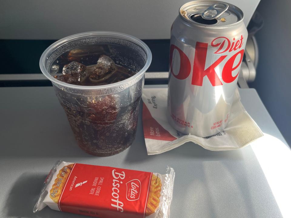 On a plane's seatback tray table, a can of Diet Coke sits on a napkin and is illuminated by a ray of sunlight. To the left of the can is a small plastic cup filled with ice and Diet Coke, and in front of the beverage is a package of Biscoff cookies.