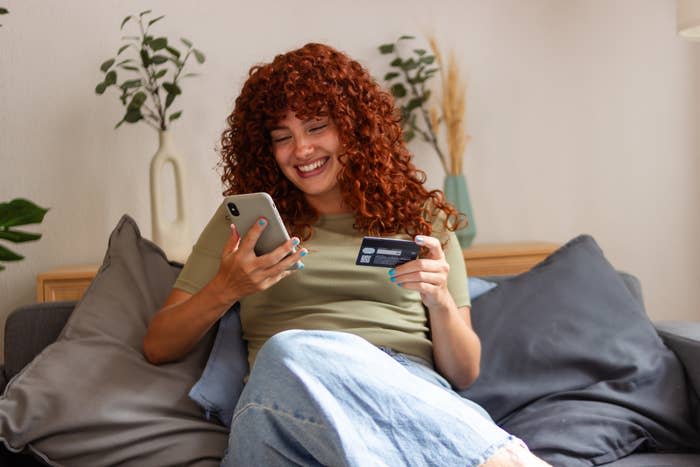 A woman with curly hair sits on a couch, smiling at her phone while holding a credit card, suggesting online shopping or mobile banking