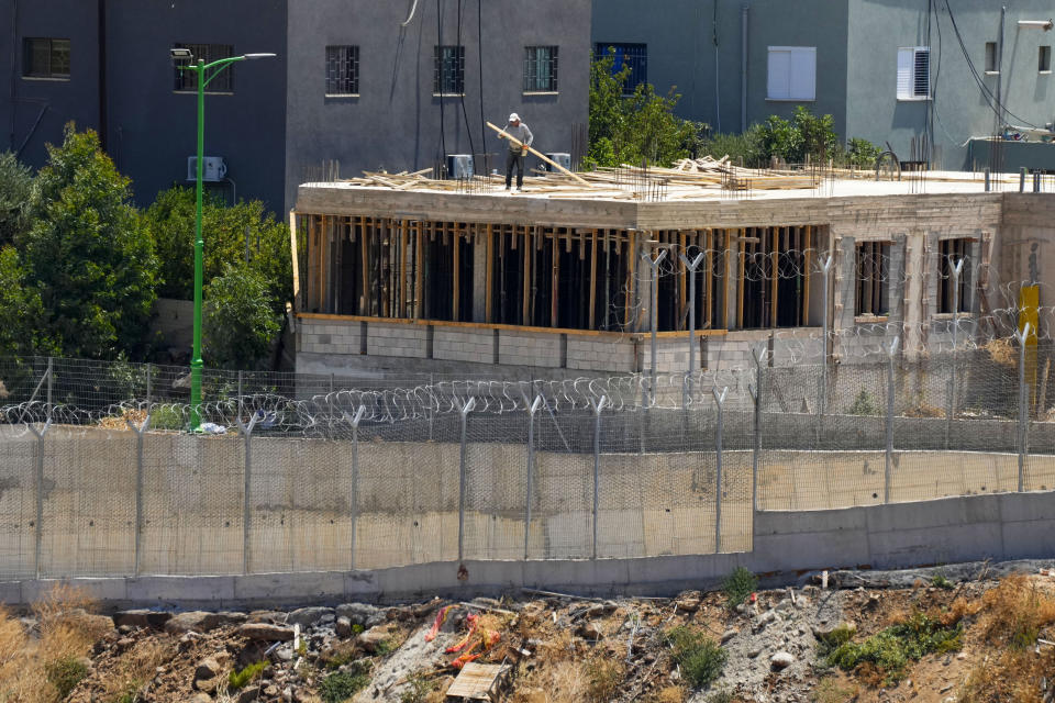 A construction worker works on the roof of a house under construction in the village of Ghajar is seen behind the newly-built wall around the Lebanese part of the village that was separated into a Lebanese section and another that is part of Syria's Israeli-occupied Golan Heights, near the Lebanese village of Wazzani, Lebanon, Tuesday, July 11, 2023. The little village of Ghajar has been a point of contention between Israel and Lebanon for years, split in two by the border between Lebanon and the Israeli-occupied Golan Heights. The dispute has begun to heat up again.(AP Photo/Hassan Ammar)