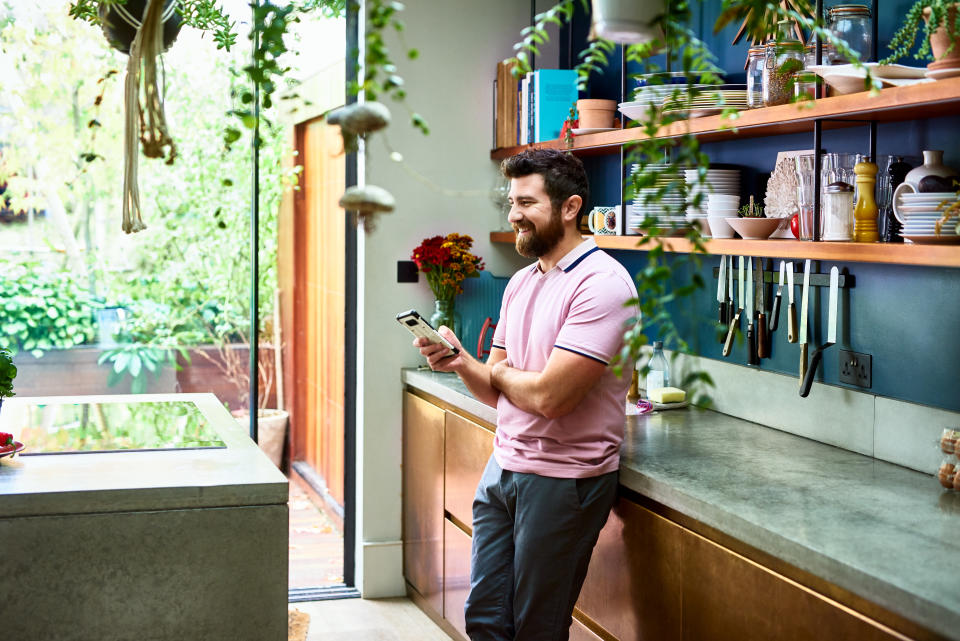 Cheerful man standing in kitchen using phone, relaxed, home interior, communication