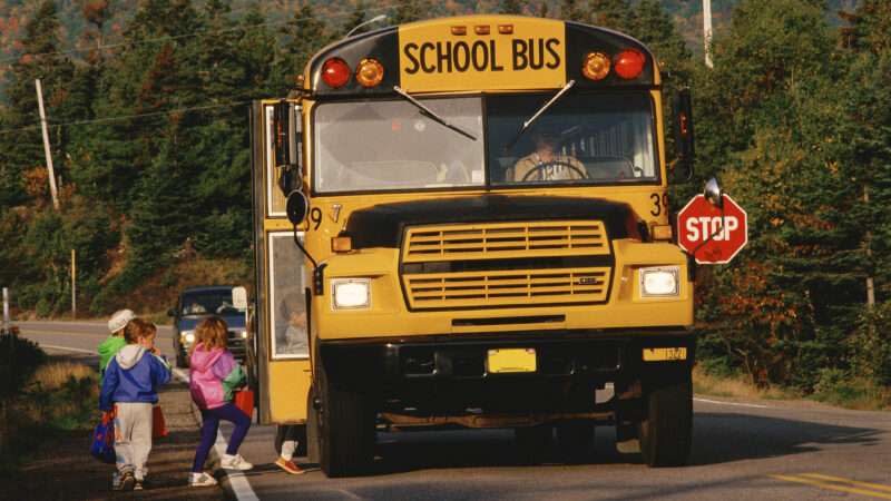 Children board a yellow school bus idling on a two-lane road.