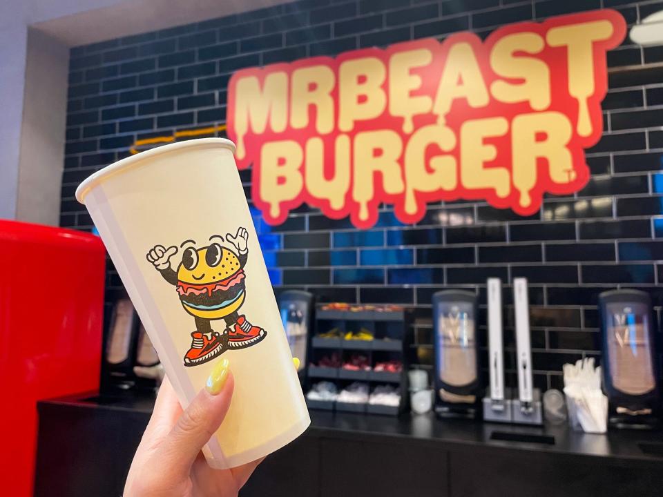 A branded cup in front of the MrBeast Burger sign