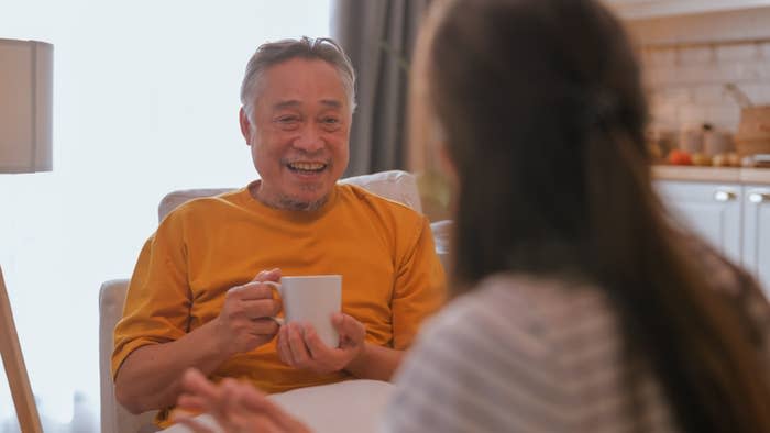 An older man with a beard smiles and holds a mug while sitting in a cozy living room, engaging in a conversation with a woman with long hair facing him