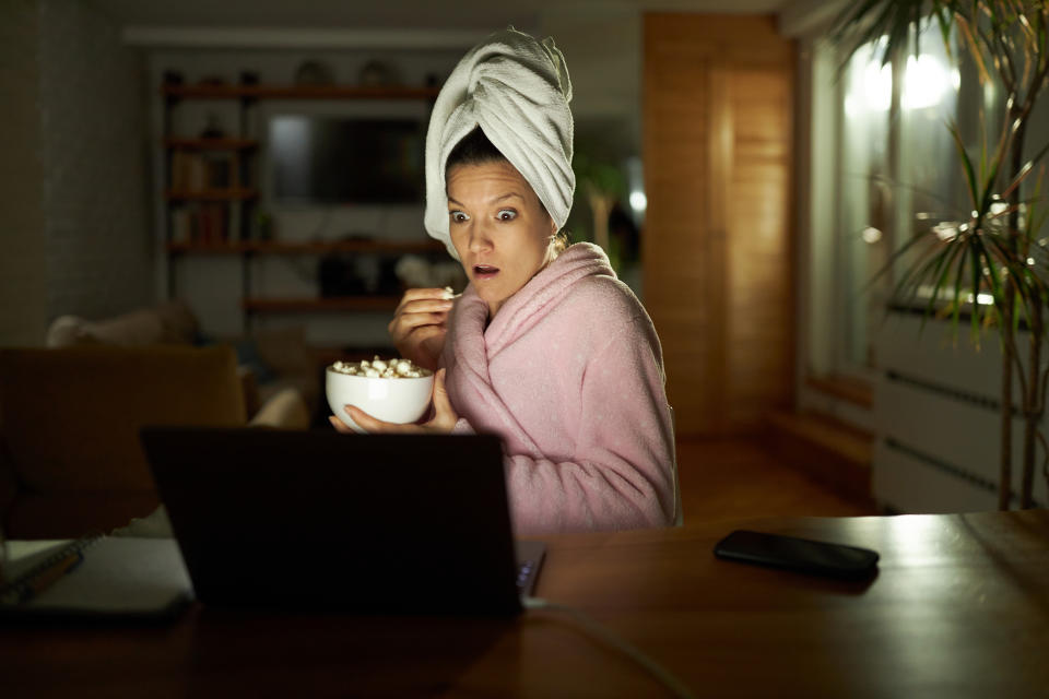 Young woman in bathrobe eating popcorn while watching a shocking movie on laptop in the evening.