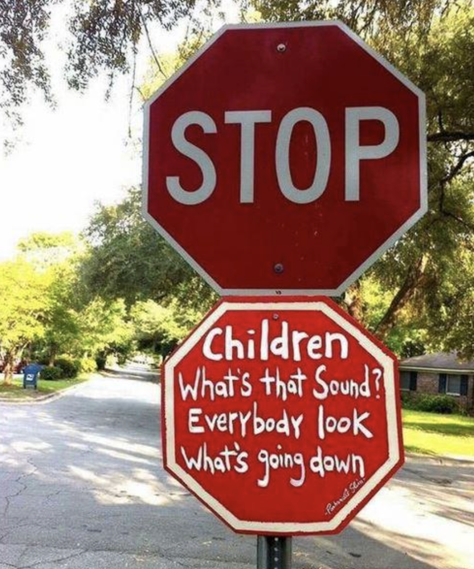 Stop sign with added text "Children What's that Sound? Everybody look What's going down" on a tree-lined street