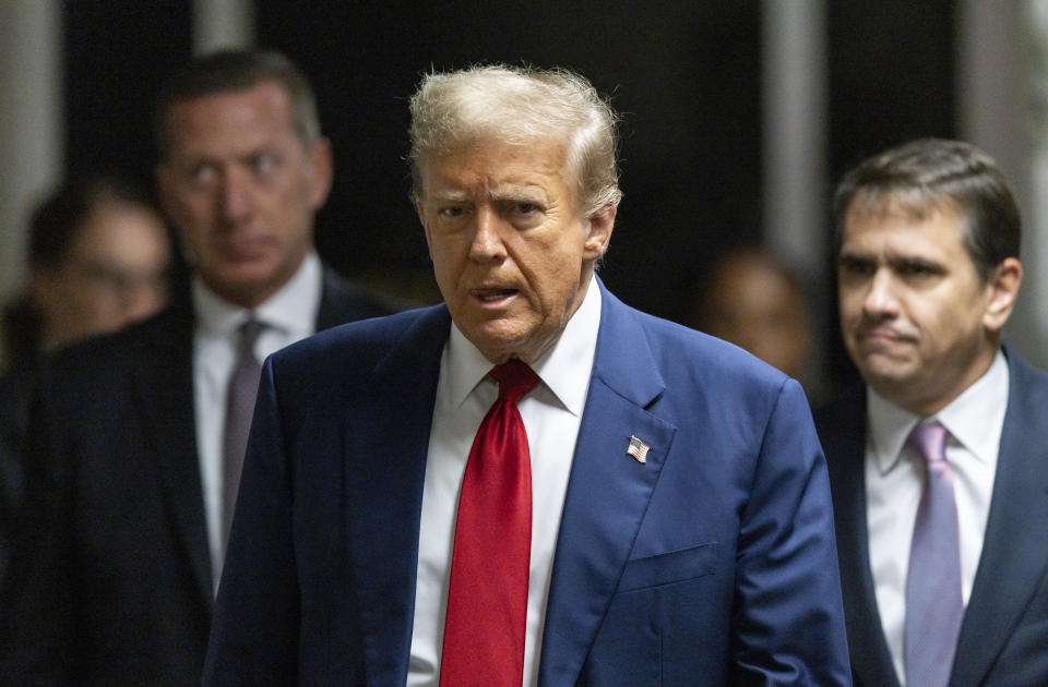 Man in suit with tie flanked by individuals in a corridor, looks serious. Expresses a newsworthy moment