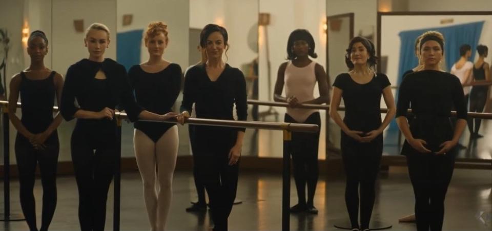 Seven women in black ballet outfits line up at the barre