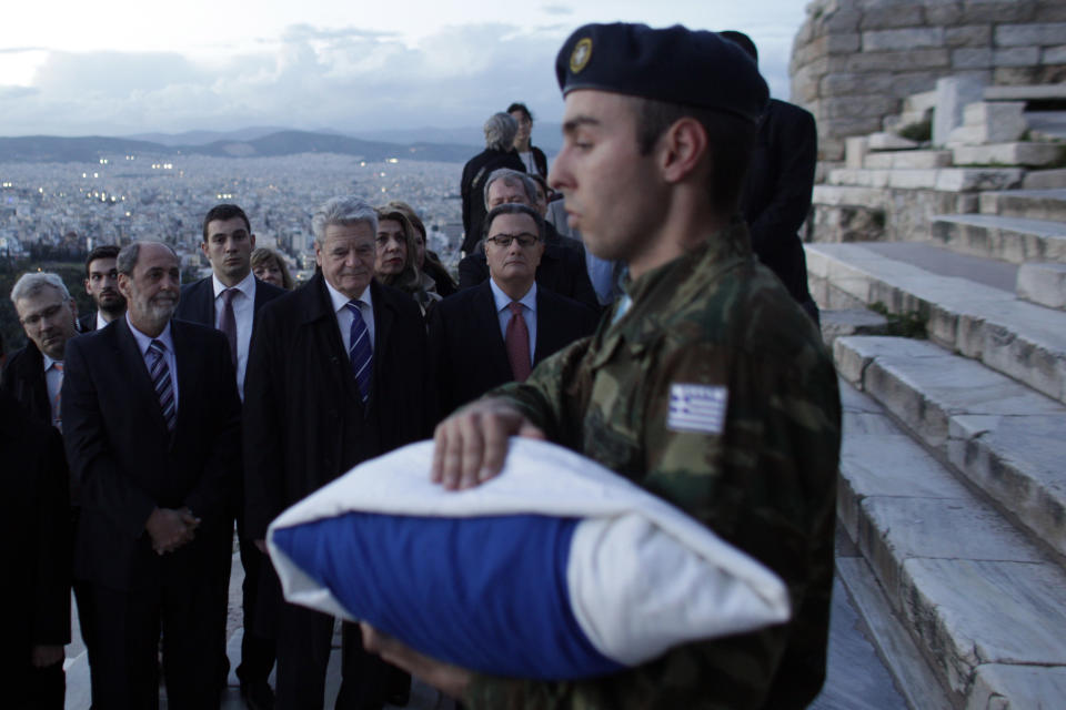 German President Joachim Gauck, center, and Greek Culture Minister Panos Panayotopoulos, right, stand to attention as a member of the presidential guard descends carrying the Greek flag that is lowered every evening from the Acropolis, on a visit to the ancient citadel in Athens, on Wednesday, March 5, 2014. Gauck is on a visit that will seek to lay to rest some of the ghosts of a brutal Nazi occupation, amid renewed anti-German sentiment stoked by Greece's financial crisis. His three-day visit will include a speech Friday at a site where German army troops massacred 92 villagers near the northeastern town of Ioannina, and a meeting with the town's Jewish community. (AP Photo/Kostas Tsironis)