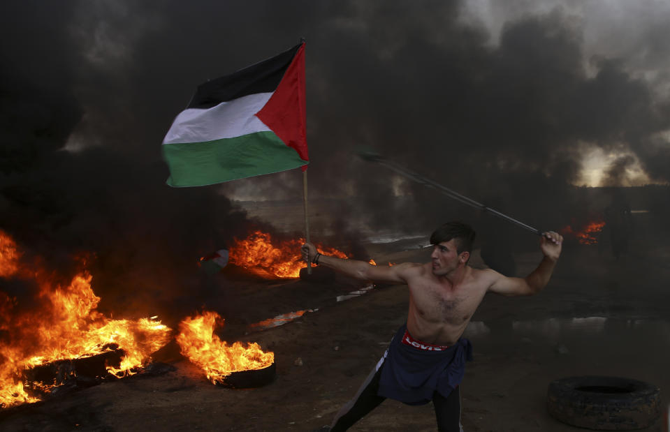 FILE-- In this Friday, Oct. 26, 2018 file photo, a protester hurls stones while holding the Palestinian flag amid burning tires near the fence of the Gaza Strip border with Israel during a protest. Palestinians in the Gaza Strip have coped with shortages of just about everything in more than a decade of border closures -- from chocolate to medicines to fuel and building supplies. Now, the past six months of protests against an Israeli-Egyptian blockade have added an unexpected item to the list of those in short supply: car tires. (AP Photo/Adel Hana, File)