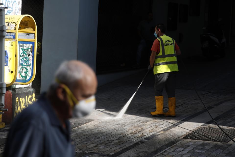 A municipal worker sprays disinfectant on a pavement as a man wearing face mask to prevent the spread of the new coronavirus walks at Ermou Street, Athens' main shopping area, Monday, Sept. 21, 2020. Greece is tightening restrictions in the greater Athens region, stepping up testing and creating quarantine hotels due to an increase in COVID-19 infections after Athenians returned from their summer holidays. (AP Photo/Thanassis Stavrakis)