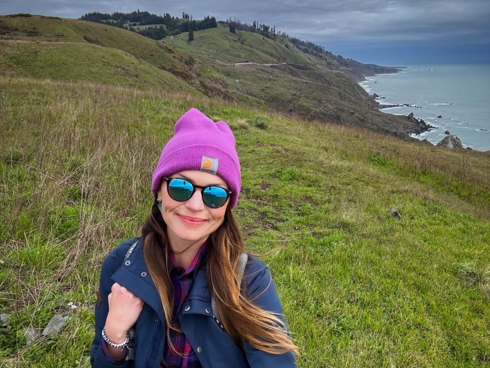 Emily, wearing a purple hat, sunglasses, a flannel, and a jacket, stands on a grassy coastline.