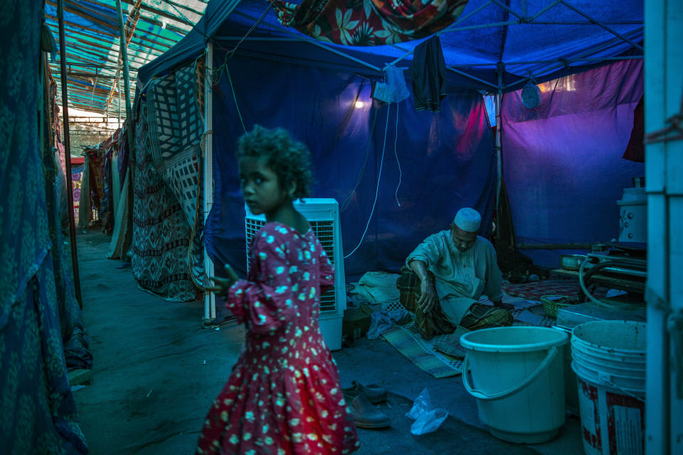 A young Rohingya refugee girl walks past an elderly man sitting inside a tent at a refugee camp alongside the banks of the Yamuna River in the south-eastern borders of New Delhi, sprawling Indian capital, July 1, 2021. Millions of refugees living in crowded camps are waiting for their COVID-19 vaccines. For months, the World Health Organization urged countries to prioritize immunizing refugees, placing them in the second priority group for at-risk people, alongside those with serious health conditions. (AP Photo/Altaf Qadri)