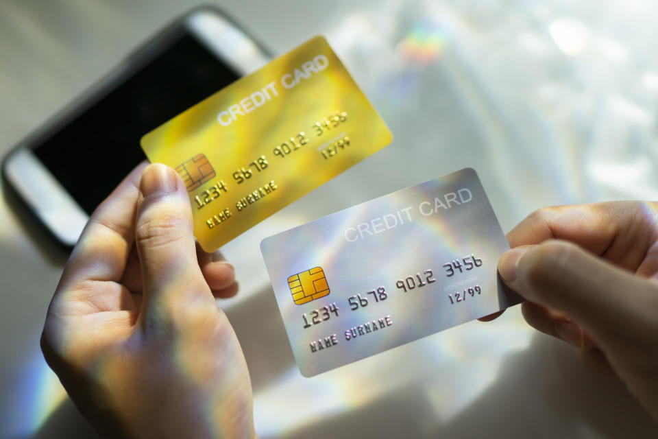 Hand holding two credit cards, one yellow and one holographic, with a smartphone nearby. The cards display generic names and numbers