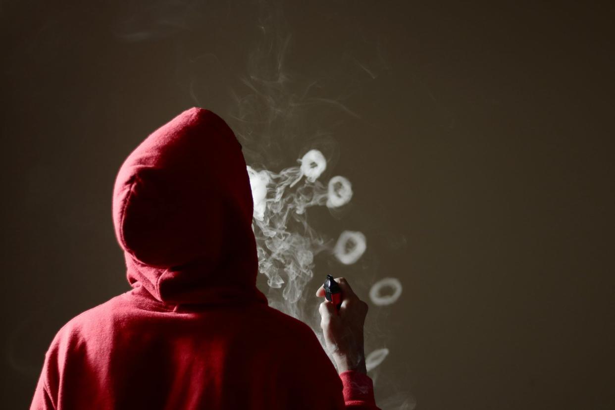 Young male in red hoodie vaping smoking, blows multiple smoke rings while holding a vape in hand, isolated rear view