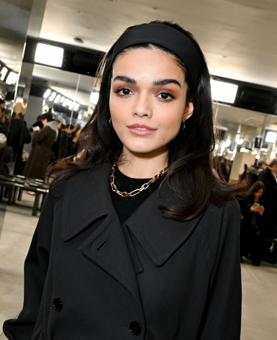 Rachel Zegler in a black trench coat with a headband, posing indoors