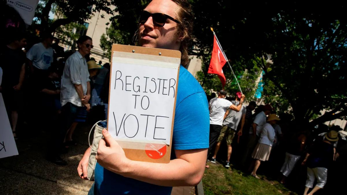 A white person wearing sunglasses holds a clipboard that says REGISTER TO VOTE on a piece of paper. A crowd of people, some holding flags, can be seen in the background. 