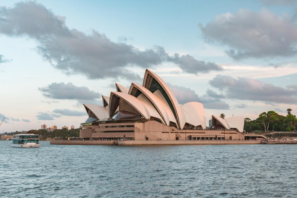 Sydney Opera House with large white sail-shaped roofs on the waterfront, under a partly cloudy sky, with a ferry boat moving in the water near it