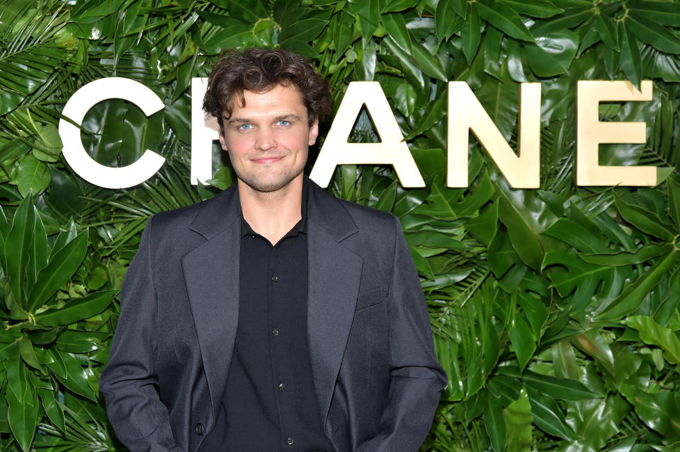 Smiling Ray in a shirt and suit jacket stands behind a leafy backdrop with "Chanel" logo