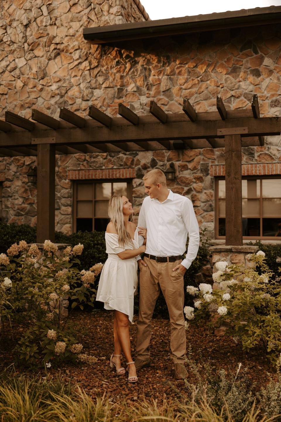 A woman in a white dress and a man in a white shirt and khakis look at each other in front of a stone building.