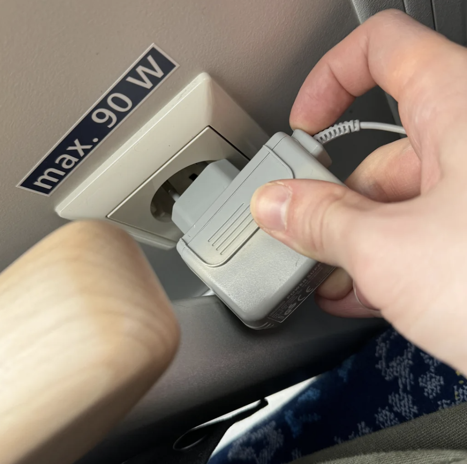 Hand plugging in a charger into an airplane seat power outlet, next to a maximum power label