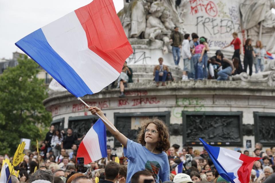 A child waving a French flag among a crowd of protesters, with people climbing a graffitied monument in the background.
