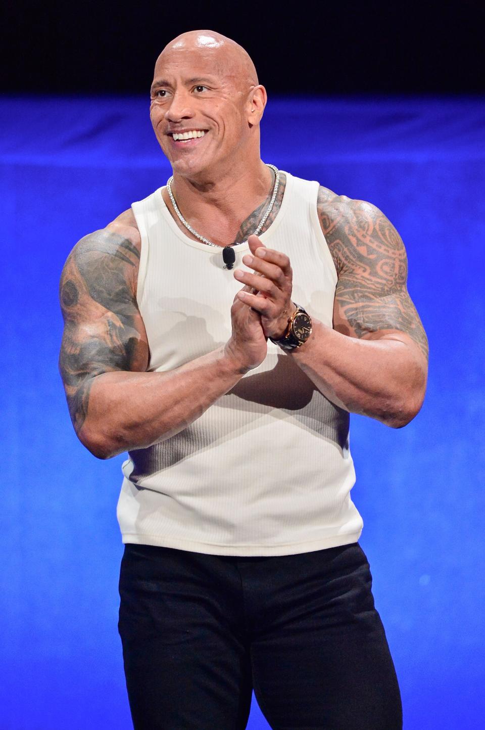 Dwayne Johnson smiles, wearing a white tank top and black pants, showcasing tattoos while clasping hands