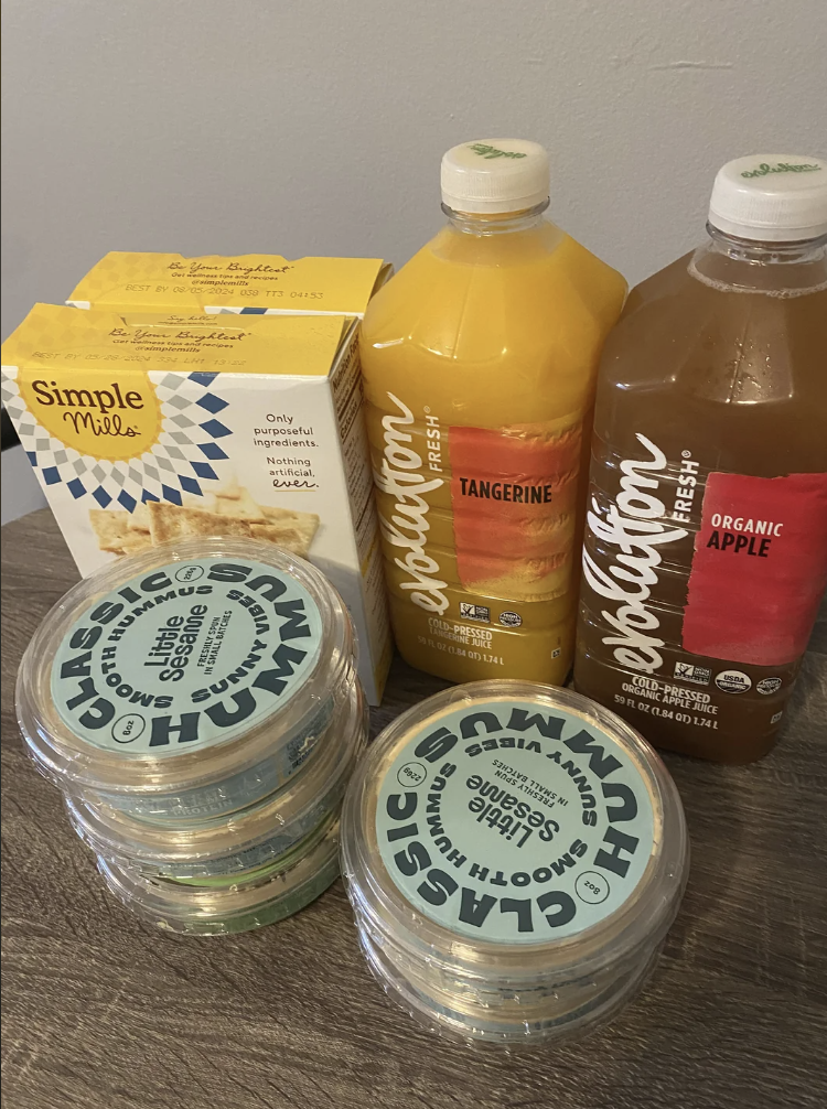 Boxes of Simple Mills crackers and bottles of Bolthouse Farms Tangerine and Organic Apple juice are on a table with four containers of Classic Hummus from Good & Gather