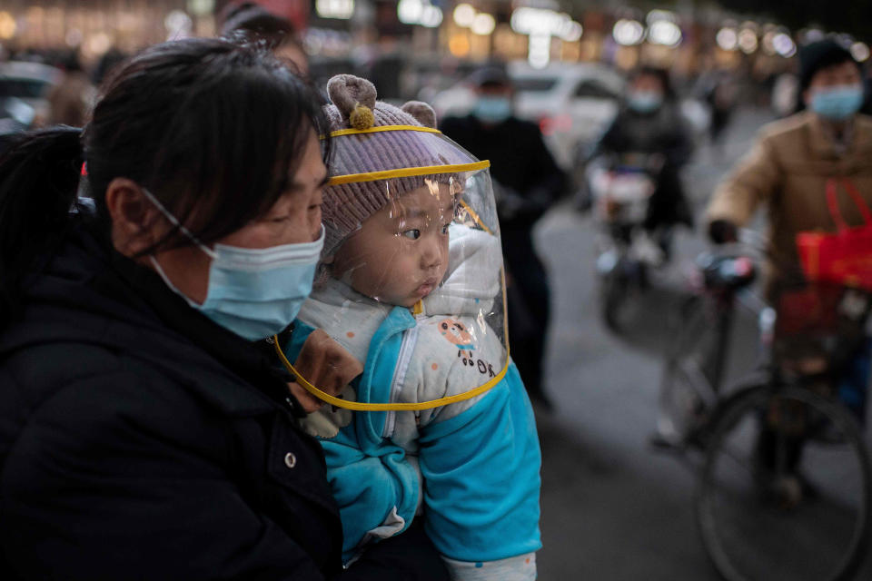 Image: A woman wearing a face mask holds a baby that wears a protective shield during rush hour on a street outside of a shopping mall complex in Wuhan  (Nicolas Asfouri / AFP - Getty Images)