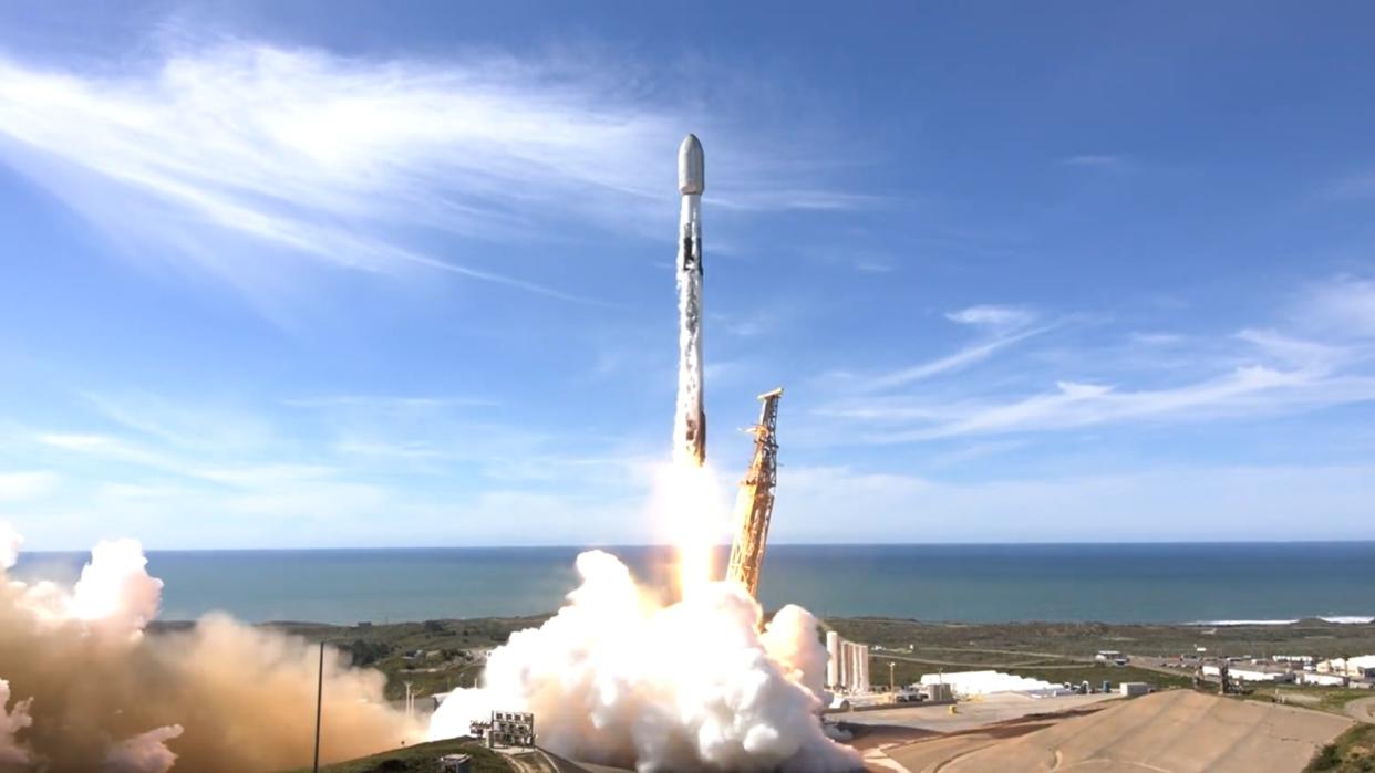  A white rocket lifts off during the day above a plume of fire. 