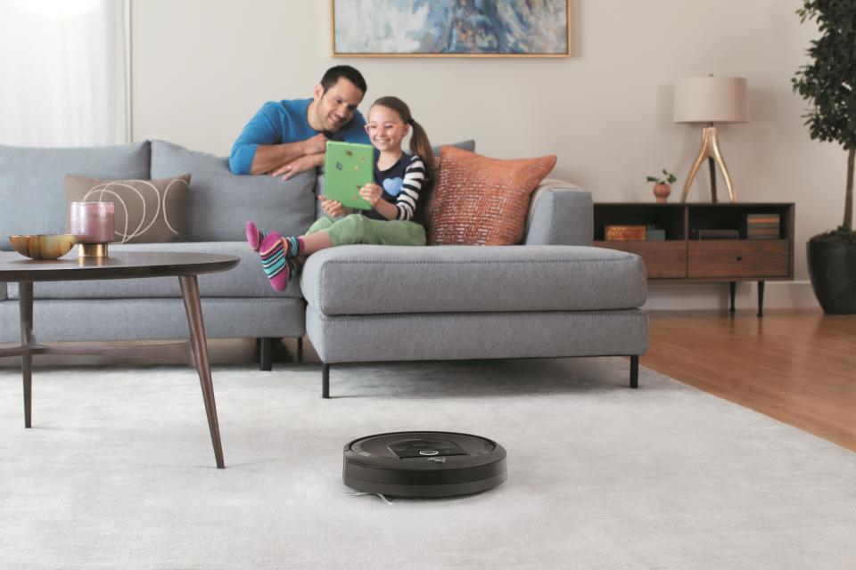 A father and young daughter sit looking at a notebook computer as a robotic vacuum cleans in the foreground.