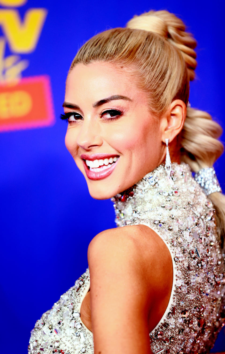 A woman on the red carpet, smiling and turning her head slightly back, wearing a sequined, high-neck dress and a high, intricately styled ponytail