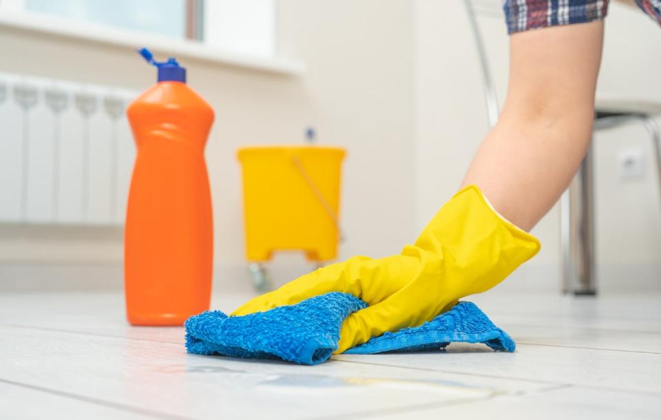 Person wearing yellow rubber gloves cleaning a white tile floor with a blue cloth; bucket and cleaning supplies in background. 