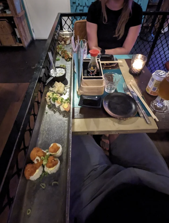 Two people dine at a restaurant with food served on a long tray between them. Various dishes and a beer are on the table