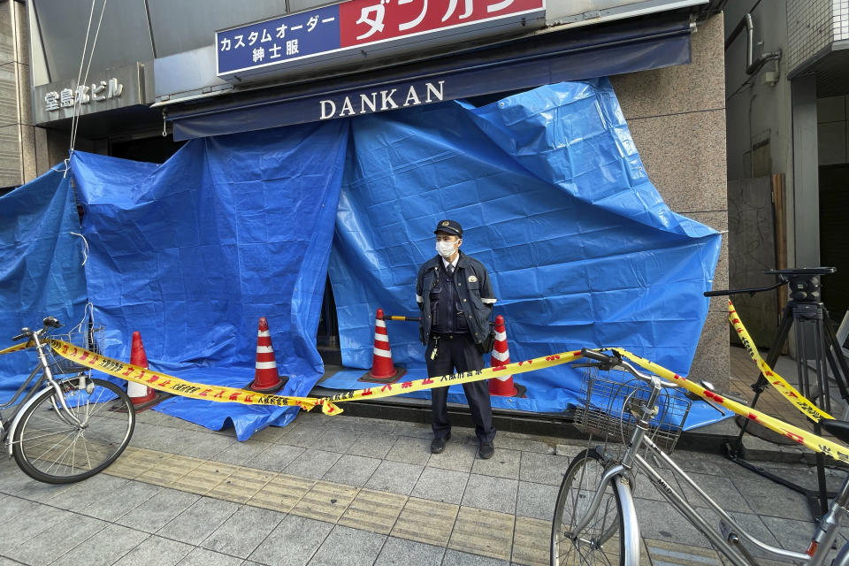 A police officer stands in front of the building where a fire broke out Friday in Osaka, western Japan, Saturday, Dec. 18, 2021. Japanese police on Saturday searched the house of one of the patients at a mental clinic where the fire gutted an entire floor in the eight-story building, killing over 20 people trapped inside. (AP Photo/Chisato Tanaka)