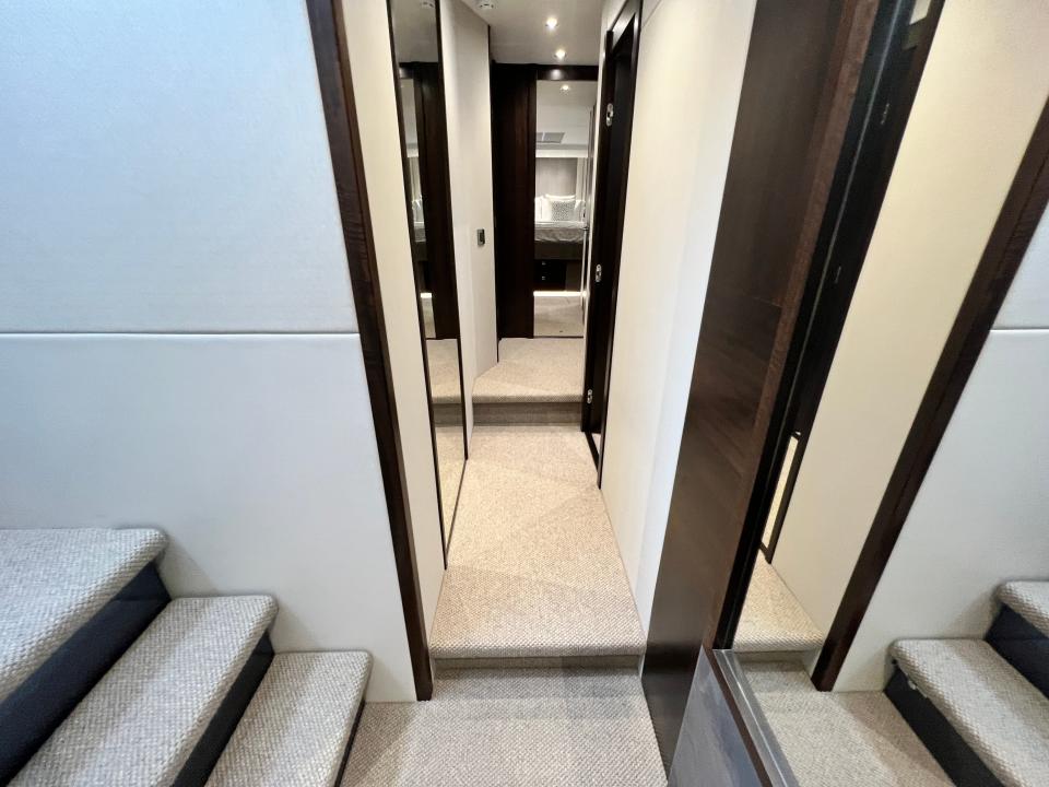 The corridor of a Sunseeker 76 yacht with grey carpets. Bedroom visible at the end, and a staircase reflected in the mirror.