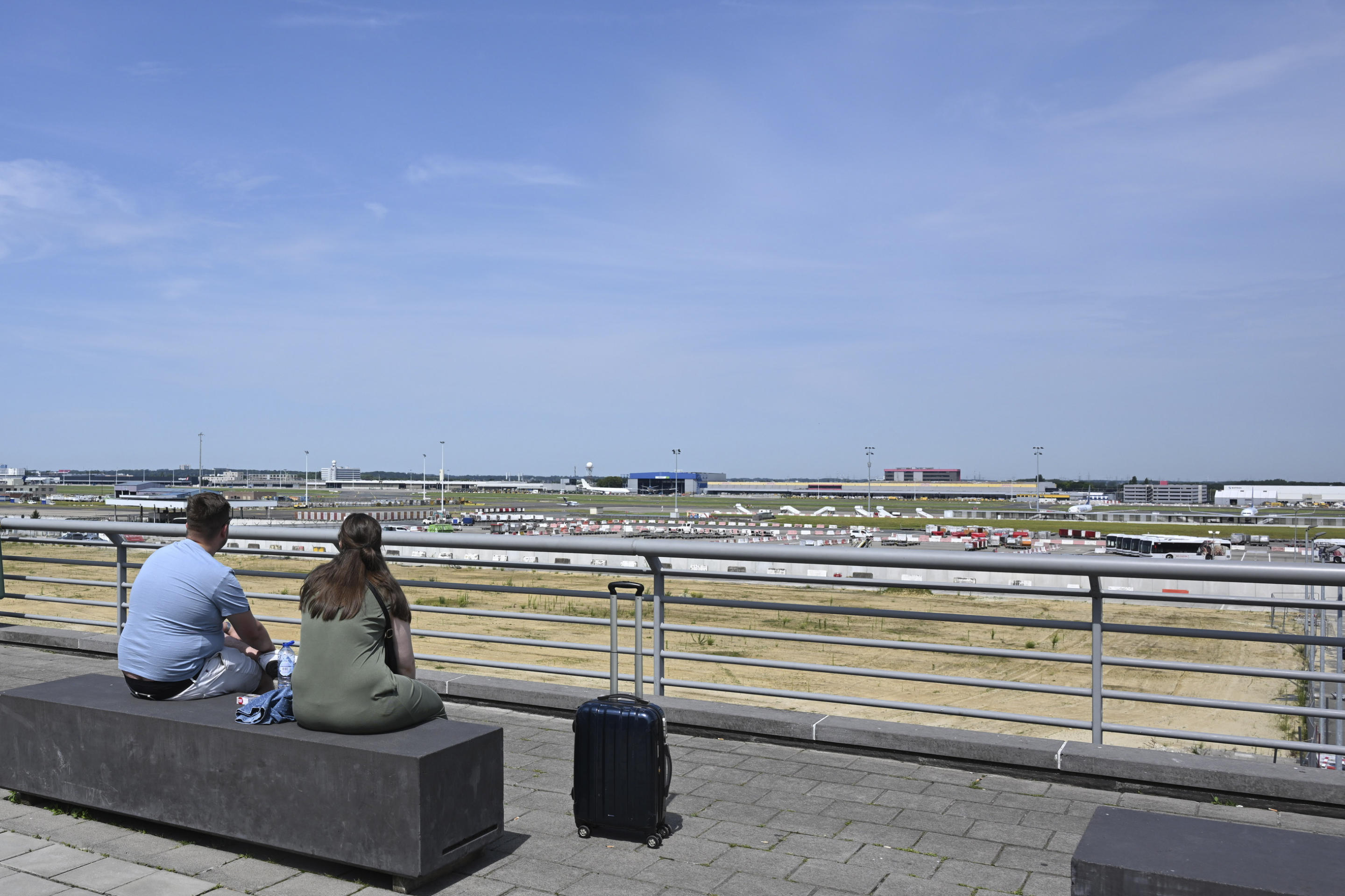 Two travelers sit on a bench overlooking the runway at Brussels airport.