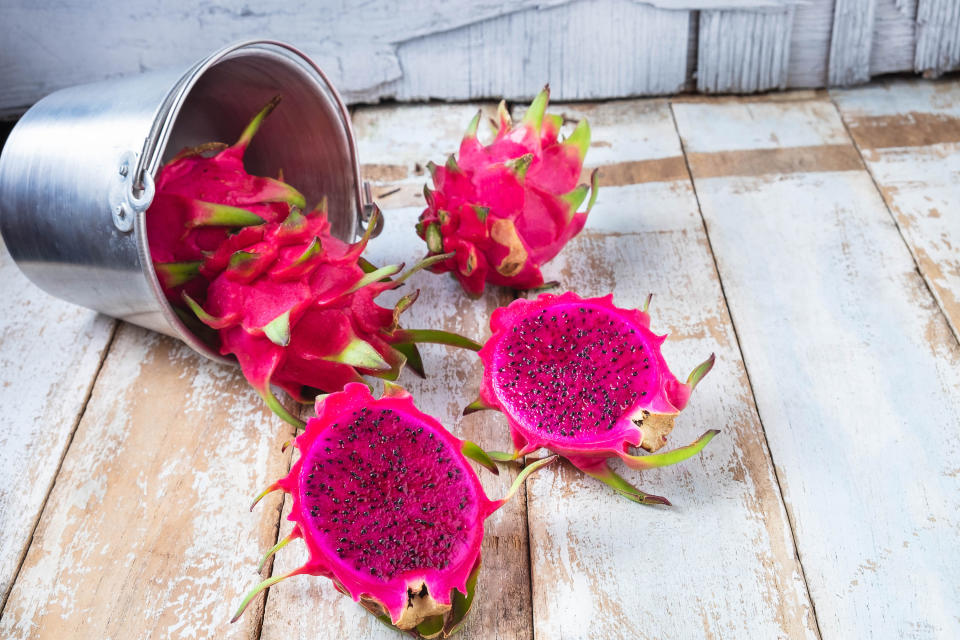 One dragon fruit is in a metal bucket tilted to the side. Two dragon fruits are on wood, and one dragon fruit that has been cut in half is also on the wood