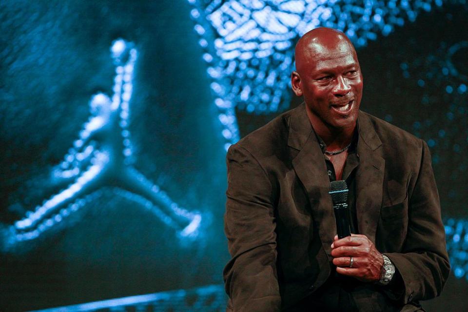 michael jordan wearing a black suit, speaking into a microphone, sitting in front of a blue air jordan logo