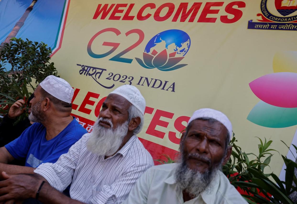 Three men sit in front of a sign advertising the G20 Summit in New Delhi, India, on Tuesday. A summit-related dinner invite that swaps in the word 'Bharat' for 'India' has triggered debate in the country. (Adnan Abidi/Reuters - image credit)