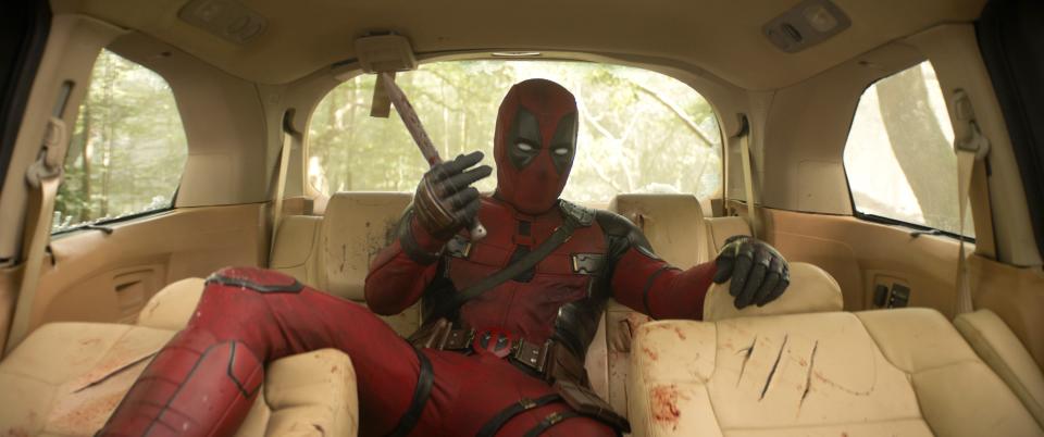 Deadpool in costume sitting in a car with one hand on the steering wheel and the other holding a katana