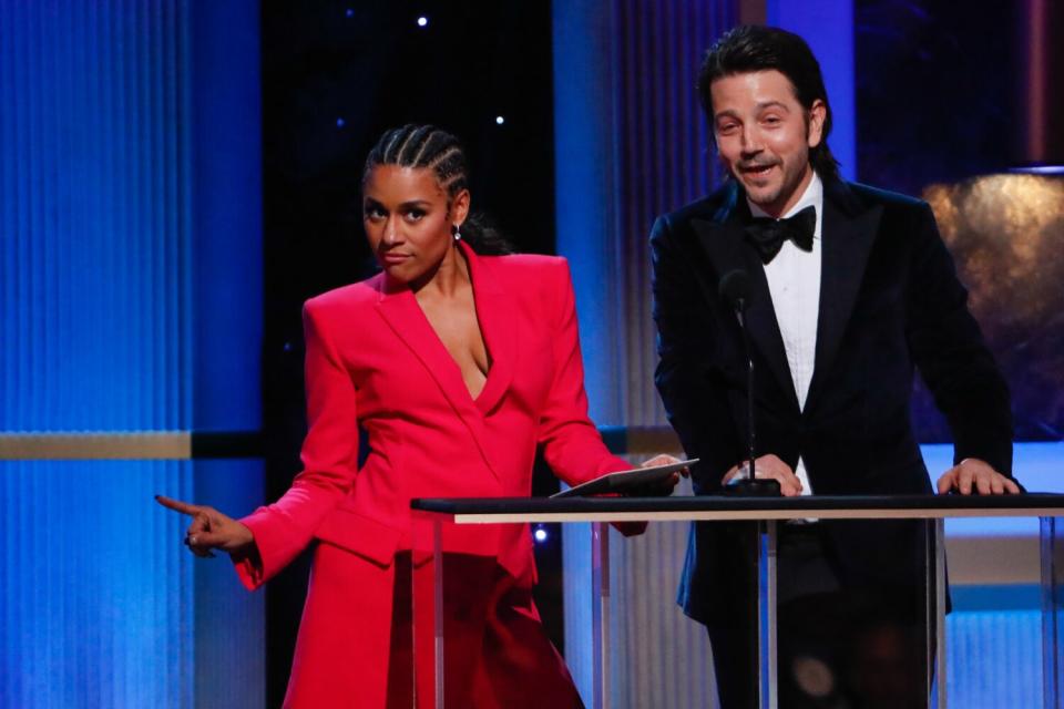 A woman posing in a pink suit and a man smiling in a black suit in front of a podium on a stage.