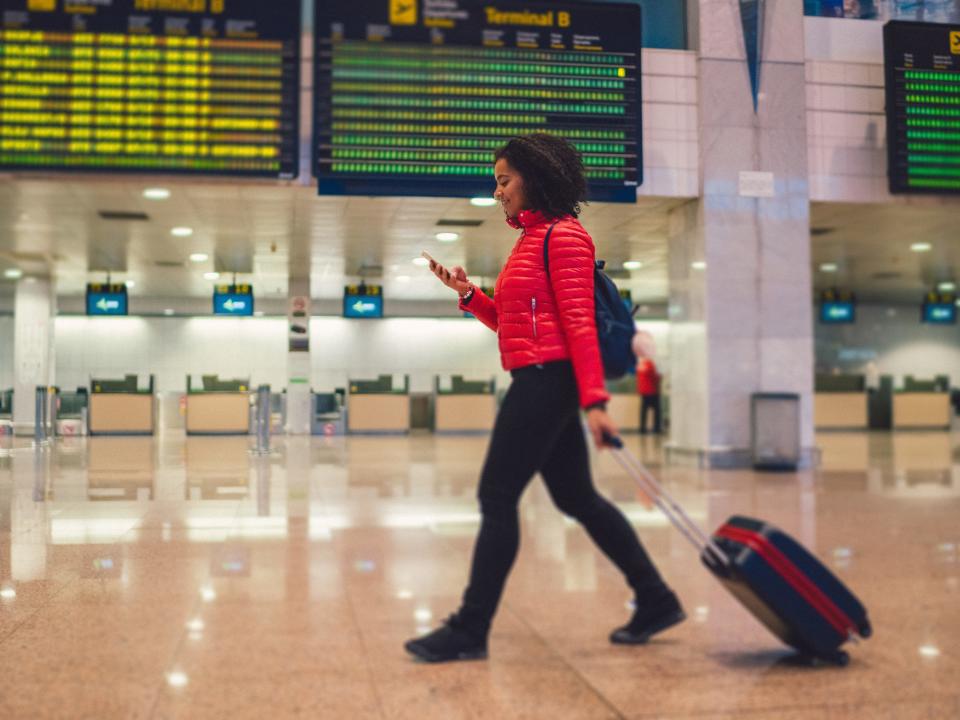 A woman rolling a suitcase through an airport, looking at her phone, in front of a departure board.