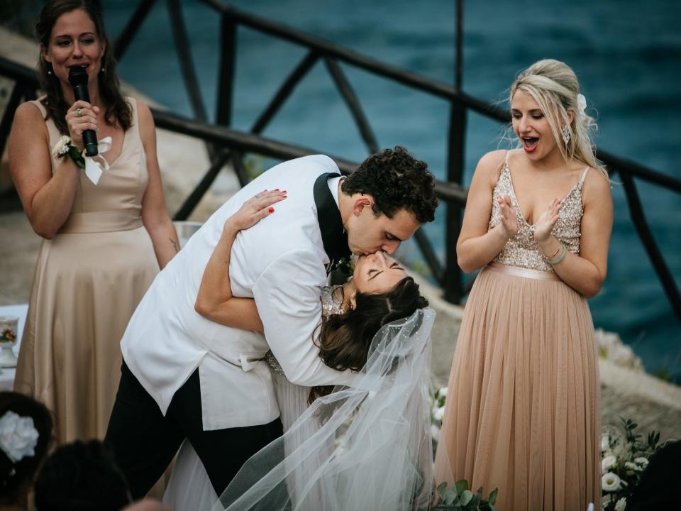 A groom dips and kisses a bride while two bridesmaids cheer.