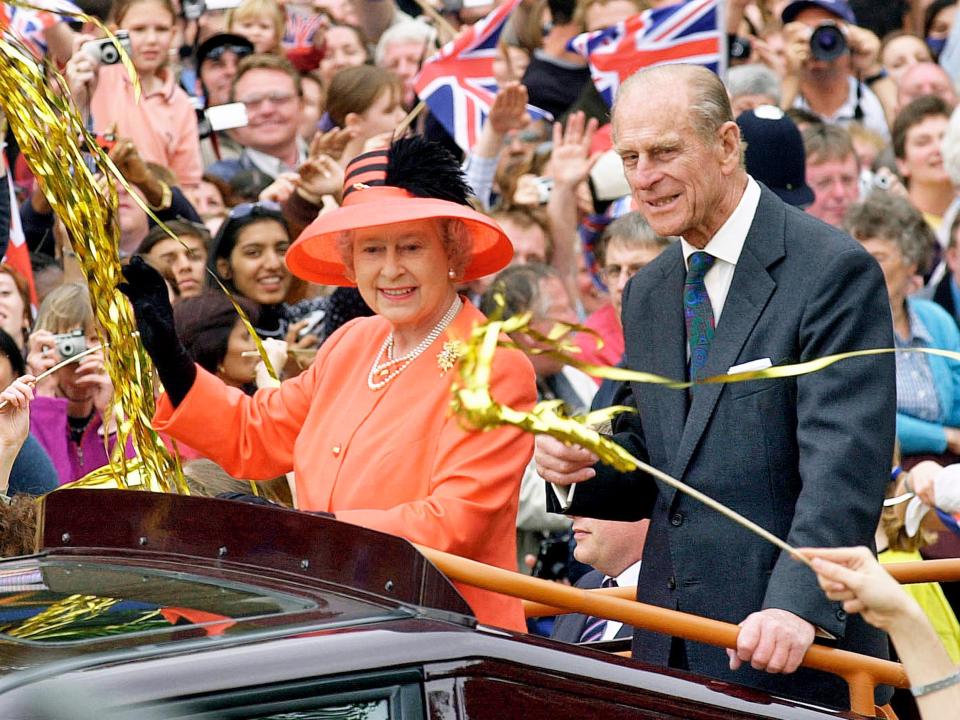 The Queen smiling and waving in an open top car surrounded by crowds. She's wearing an orange overcoat with gold buttons. She has a necklace with layers of pearls, a wide-brimmed orange hat with black stripes and a black feather, and black gloves.